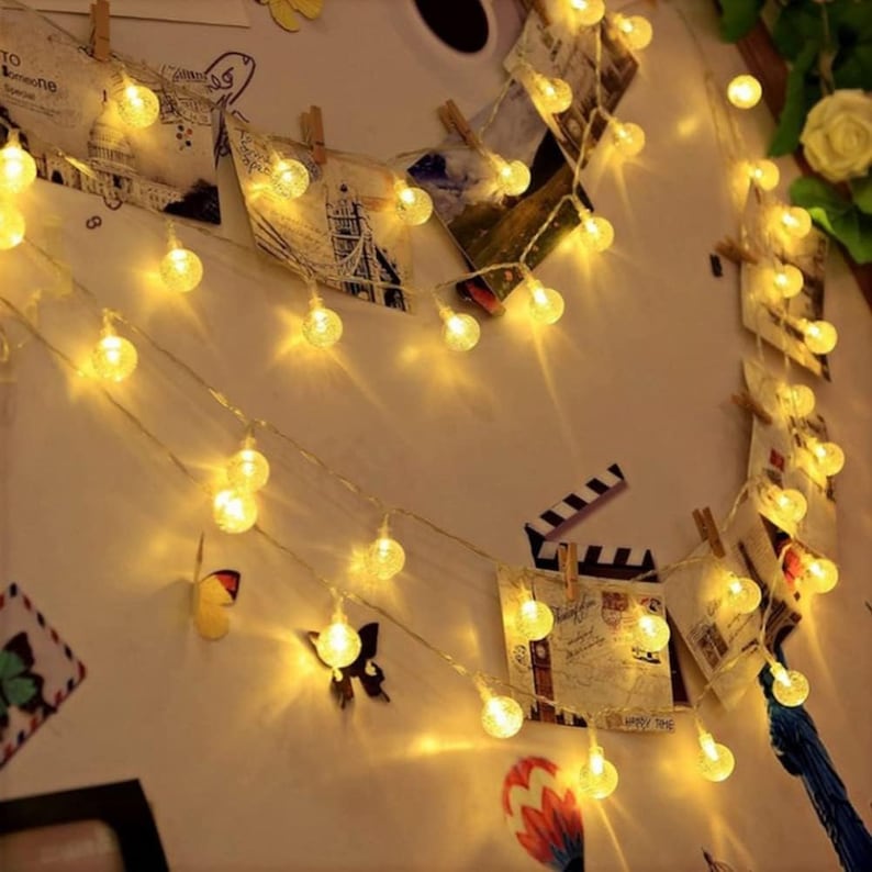 Puede incluir: Luces de cadena con peque&ntilde;as bombillas redondas y brillantes, de color amarillo c&aacute;lido, colgadas en una pared. Las luces se intercalan con peque&ntilde;as fotos y recortes decorativos. El fondo es de color beige claro.