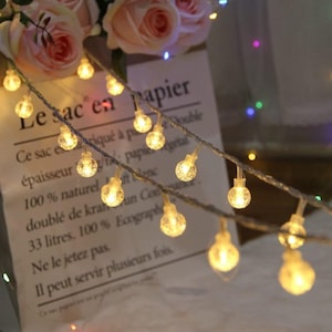 Puede incluir: Cadena de peque&ntilde;as luces LED redondas y transparentes, que parecen peque&ntilde;as burbujas, ensartadas en un cable plateado. Las luces emiten un brillo c&aacute;lido y amarillo. El fondo incluye un ramo de rosas rosas y un papel con texto en franc&eacute;s.