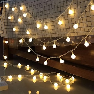 Puede incluir: Cadena de luces con bombillas esf&eacute;ricas blancas c&aacute;lidas. Las luces est&aacute;n ensartadas en un cable transparente y est&aacute;n colgadas sobre una estructura de cama y una tela estampada. Las luces est&aacute;n espaciadas uniformemente y emiten un brillo suave.