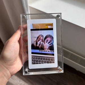May include: A clear acrylic photo frame with a white mount and a photo of two people covering their faces with their hands. The photo is taken in front of a laptop with a keyboard.