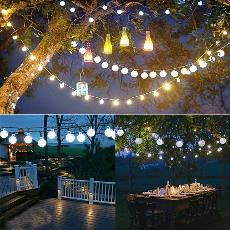 Puede incluir: Luces de cadena para exteriores con bombillas redondas blancas y un brillo blanco c&aacute;lido, colgadas de las ramas de los &aacute;rboles. La imagen tambi&eacute;n muestra botellas de vidrio decorativas y una linterna colgando del &aacute;rbol, creando un ambiente festivo.