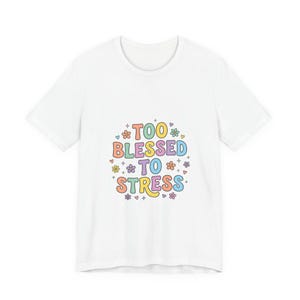 Op de afbeelding: Een wit t-shirt met korte mouwen en de tekst "TOO BLESSED TO STRESS" in een veelkleurig, afgerond lettertype. Het ontwerp is omringd door kleine, kleurrijke bloem- en hartpictogrammen in tinten roze, geel, blauw, oranje en paars.