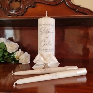 May include: Two white taper candles with the names "Siobhan" and "Cedlan" inscribed on them. A larger white pillar candle with the text "The Wedding of Siobhan and Cedlan, 31st March 2023, Ten Square" is in the background.