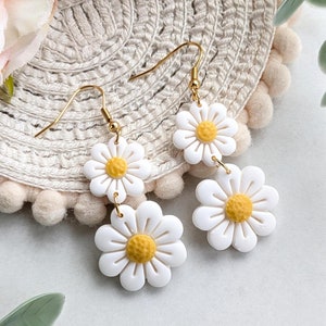 May include: A pair of gold dangle earrings with two white daisy flower charms. The daisies have yellow centers.