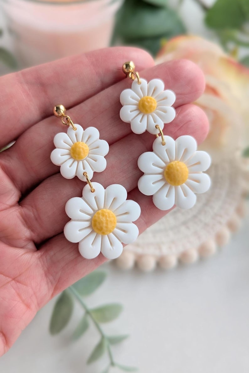 May include: A pair of white daisy earrings with yellow centers. The earrings are made of polymer clay and have gold-toned hooks.