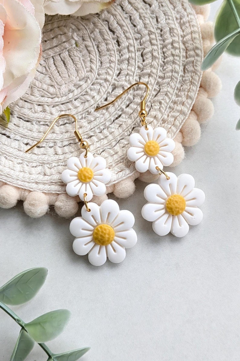 May include: A pair of gold dangle earrings with two white daisy flower charms. The daisies have yellow centers.