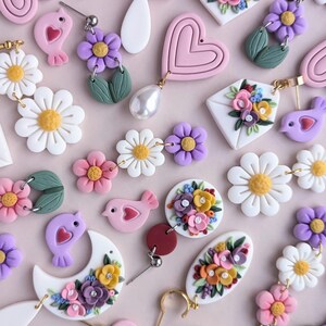 May include: Assortment of handmade polymer clay earrings featuring floral designs, hearts, birds, and daisies. The earrings are in shades of pink, purple, white, and yellow.
