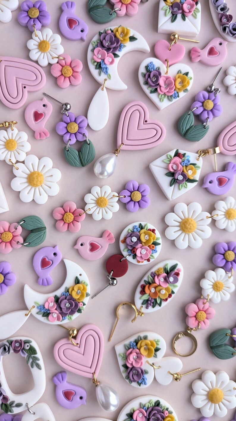 May include: Assortment of handmade polymer clay earrings featuring floral designs, hearts, birds, and daisies. The earrings are in shades of pink, purple, white, and yellow.