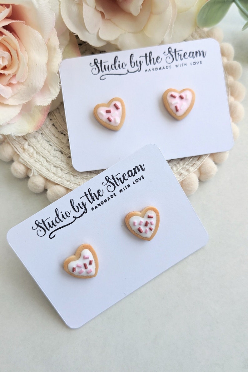 Puede incluir: Dos pares de pendientes de galleta en forma de coraz&oacute;n con chispas rosas y marrones en una tarjeta blanca con el texto "Studio by the Stream Handmade with Love".