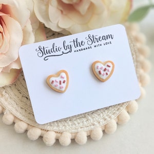 Puede incluir: Un par de pendientes en forma de coraz&oacute;n de galleta con glaseado rosa y blanco y chispas. Los pendientes est&aacute;n en una tarjeta blanca con el texto "Studio by the Stream Handmade with Love".