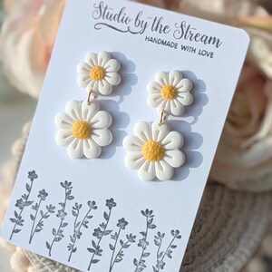 May include: A pair of white daisy earrings with yellow centers. The earrings are on a white card with the text "Studio by the Stream Handmade with Love".