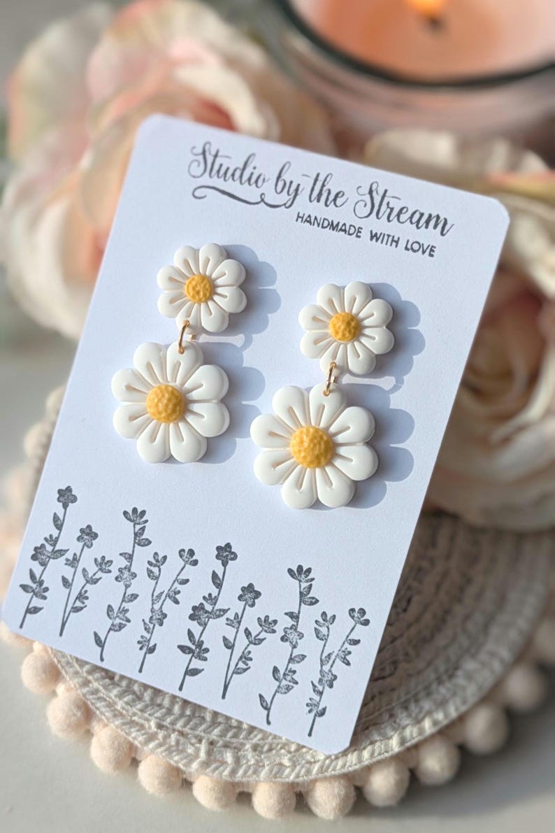 May include: A pair of white daisy earrings with yellow centers. The earrings are on a white card with the text "Studio by the Stream Handmade with Love".