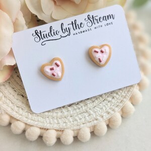Puede incluir: Un par de pendientes en forma de coraz&oacute;n de galleta con glaseado rosa y chispas rojas. Los pendientes se muestran en una tarjeta blanca con el texto "Studio by the Stream Handmade with Love".