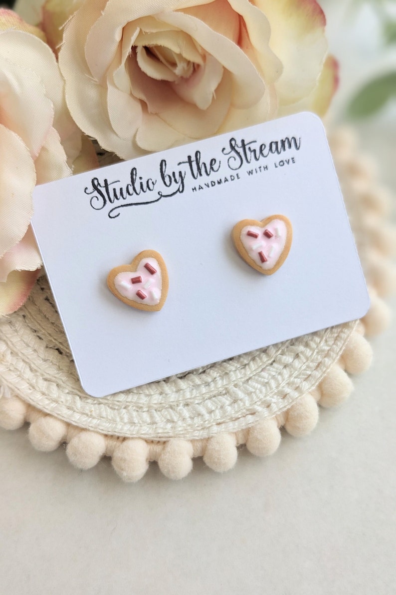 Puede incluir: Un par de pendientes en forma de coraz&oacute;n de galleta con glaseado rosa y chispas rojas. Los pendientes se muestran en una tarjeta blanca con el texto "Studio by the Stream Handmade with Love".