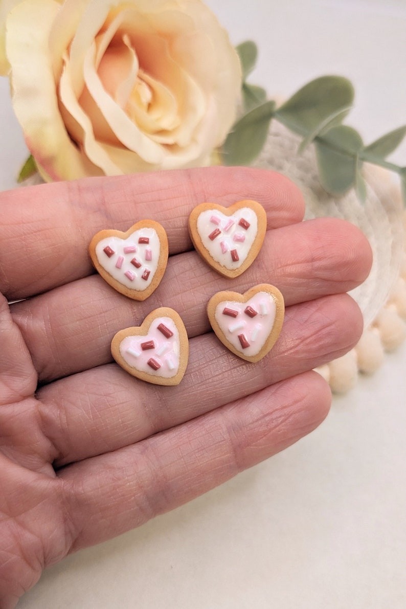Puede incluir: Cuatro galletas en forma de coraz&oacute;n con chispas rosas y marrones en la parte superior. Las galletas est&aacute;n en una mano.