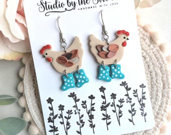 May include: A pair of white chicken earrings with blue and white polka dot rain boots. The earrings are on a white card with the text "Studio by the Stream Handmade with Love".