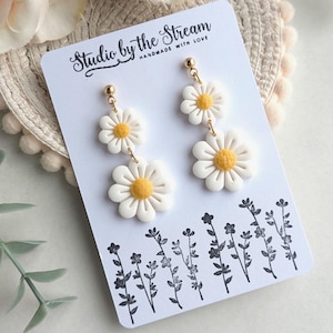 May include: A pair of white daisy earrings with yellow centers. The earrings are on a white card with the text "Studio by the Stream Handmade with Love".