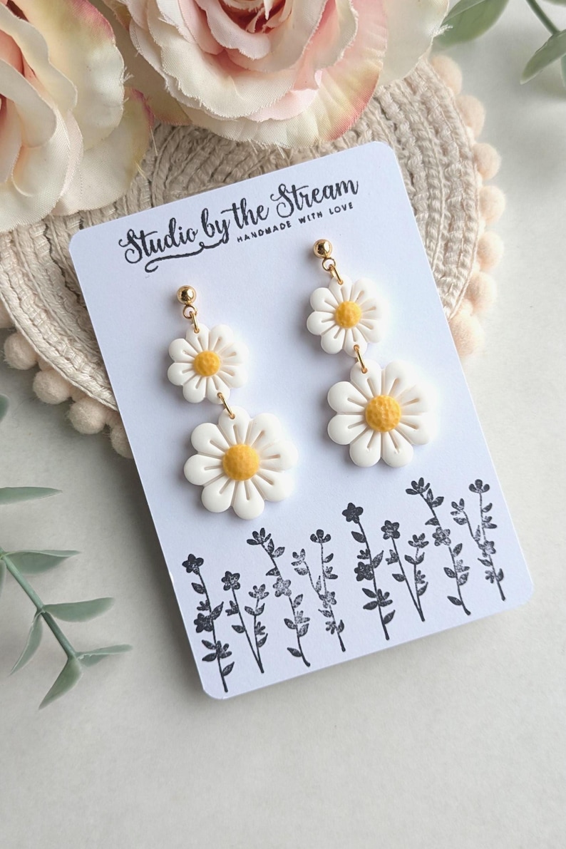 May include: A pair of white daisy earrings with yellow centers. The earrings are on a white card with the text "Studio by the Stream Handmade with Love".