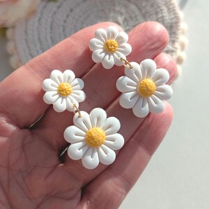 May include: A pair of white daisy earrings with yellow centers. The earrings are made of polymer clay and have a dangling design.