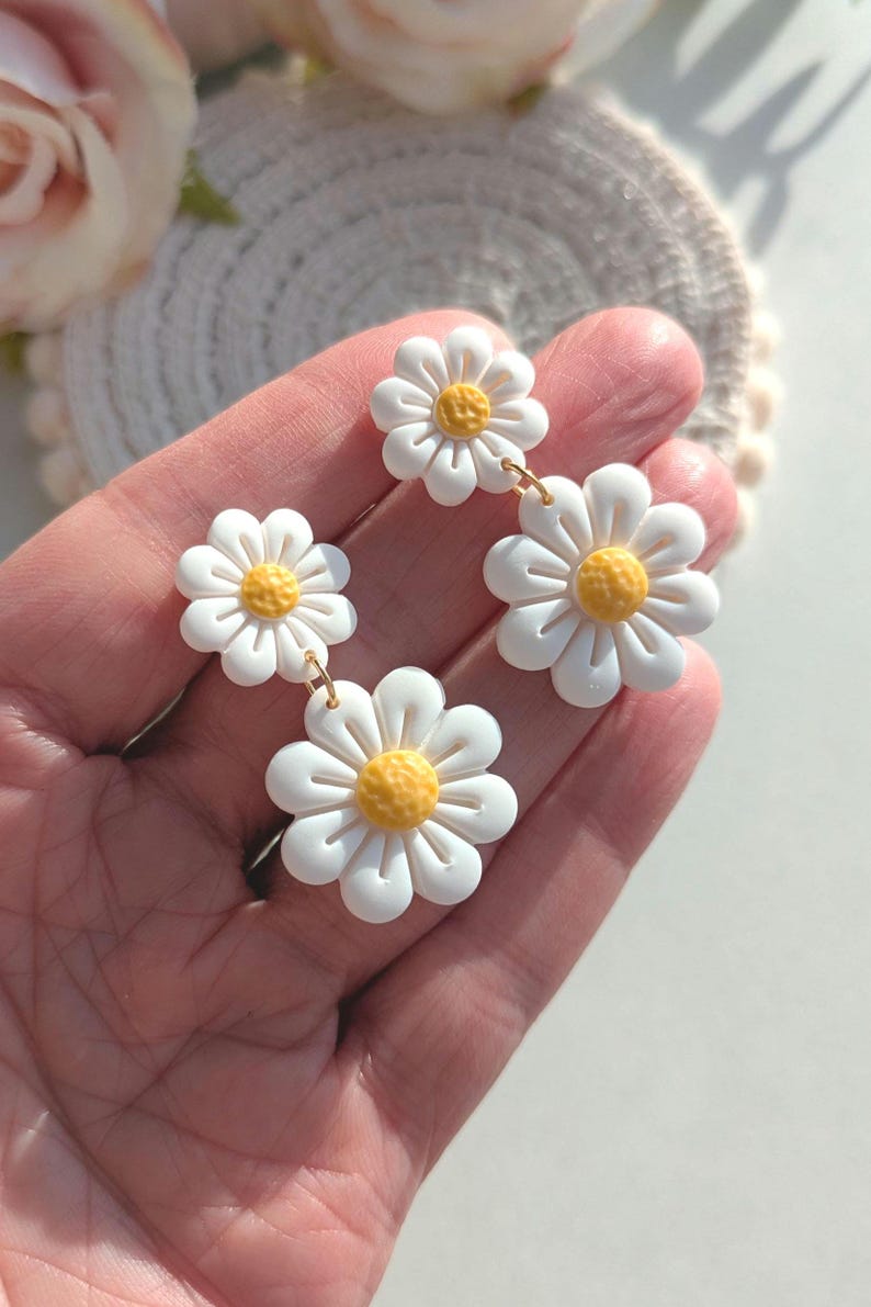 May include: A pair of white daisy earrings with yellow centers. The earrings are made of polymer clay and have a dangling design.