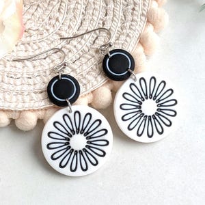 May include: A pair of dangle earrings with a black and white design. Each earring has a small black circle with a white outline above a larger white circle with a black floral pattern. The earrings hang from silver-coloured hooks.
