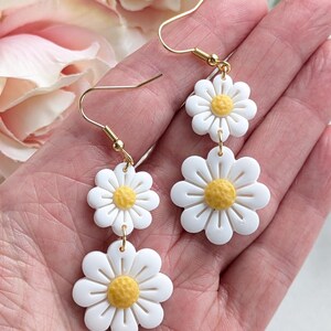 May include: A pair of gold dangle earrings with three white daisy flower charms. The center of each flower is yellow.