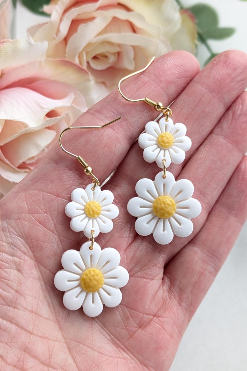 May include: A pair of gold dangle earrings with three white daisy flower charms. The center of each flower is yellow.