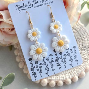May include: A pair of gold dangle earrings with three white daisy flowers on each earring. The earrings are displayed on a white card with the text "Studio by the Stream Handmade with Love".