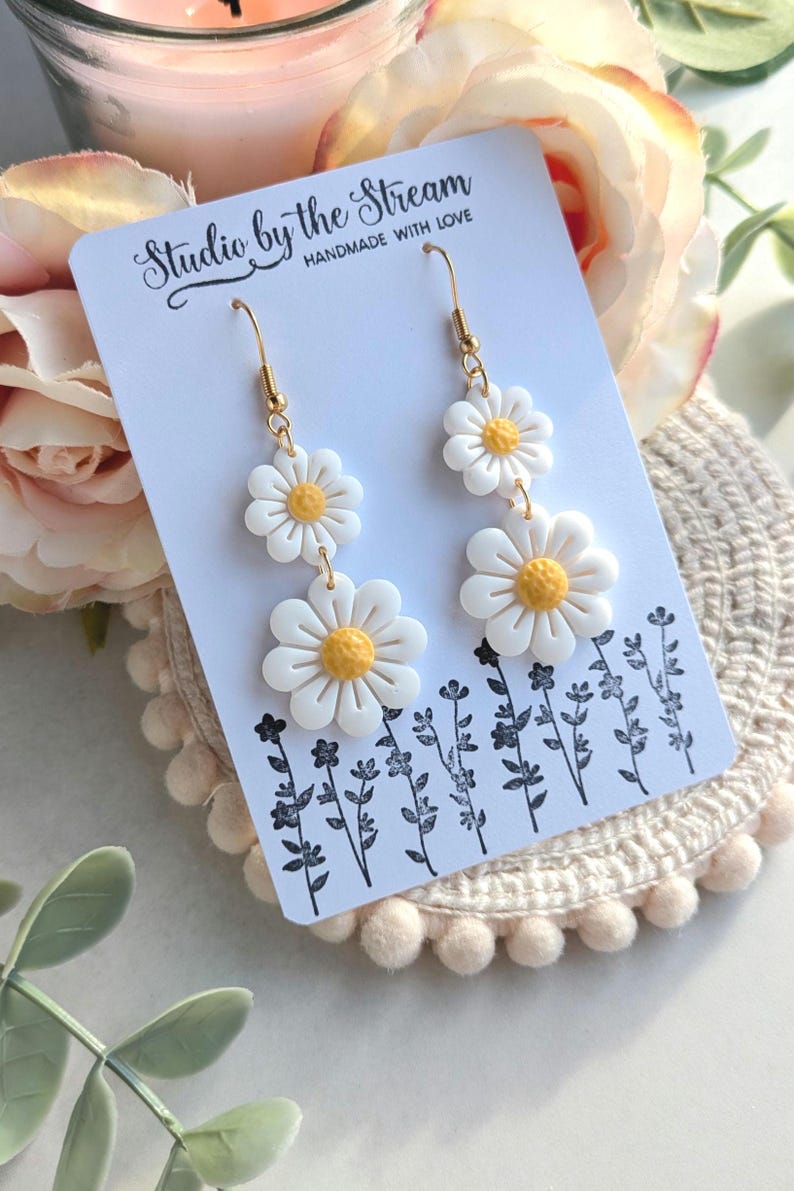 May include: A pair of gold dangle earrings with three white daisy flowers on each earring. The earrings are displayed on a white card with the text "Studio by the Stream Handmade with Love".