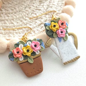 Puede incluir: Un par de pendientes hechos a mano con un tema floral. Un pendiente es una maceta marrón con flores coloridas. El otro pendiente es una regadera blanca con detalles dorados y flores coloridas. Ambos pendientes tienen ganchos dorados.
