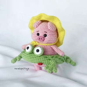 May include: A handmade crochet pig wearing a yellow hat and scarf, riding a green frog. The pig is pink with black eyes and a small snout. The frog has large white and black eyes and a red mouth. The toy is set against a white background.