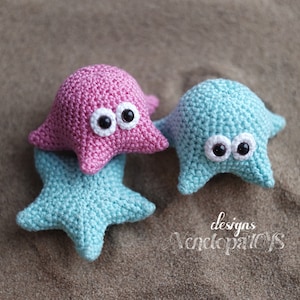 May include: Three handmade crochet starfish toys in shades of pink and blue, with white and black eye details. The toys are arranged on a sandy surface, showcasing a whimsical, beach-themed craft.