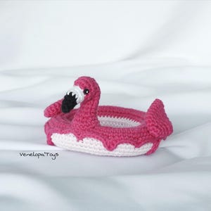 May include: A handmade crochet flamingo floatie in shades of pink and white. The flamingo has a long neck, black beak, and white accents. The floatie is designed with a circular shape and a decorative edge. The text "VenelopaToys" is visible.