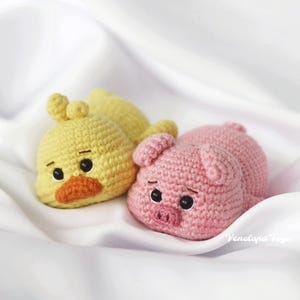 Può includere: Due animali di peluche all'uncinetto, un'anatra gialla e un maiale rosa, sdraiati su uno sfondo di tessuto bianco.