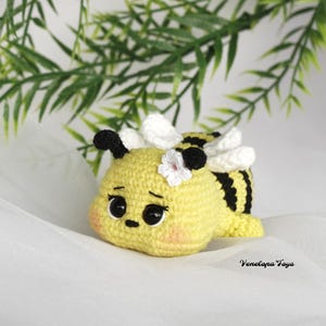 Pode incluir: Um brinquedo de abelha de crochê com listras amarelas e pretas, asas brancas e uma flor branca na cabeça. A abelha tem olhos pretos grandes e uma expressão fofa.
