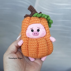 May include: A crocheted plush toy pig wearing a pumpkin costume. The pig is pink with black eyes and a small pink nose. The pumpkin costume is orange with a green stem and leaves.