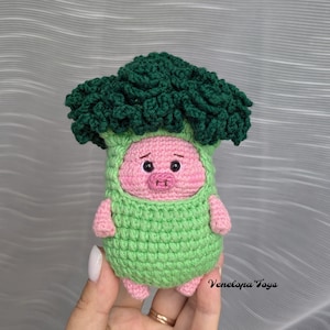 May include: A crocheted pig wearing a green broccoli costume. The pig has pink skin, black eyes, and a small pink snout. The broccoli is green and has a fluffy texture.
