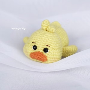 May include: A crocheted yellow chick toy with black eyes and an orange beak. The chick is lying on a white fabric surface.