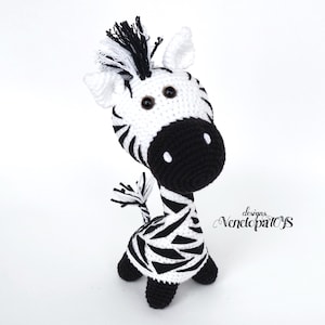 May include: A black and white crocheted zebra toy with black eyes and a black nose. The zebra is standing on a white surface.