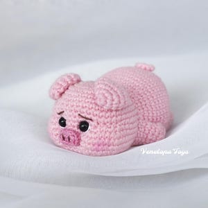 Puede incluir: Un juguete de cerdo rosa hecho a mano con ganchillo, tumbado. El cerdo tiene ojos negros, un hocico rosa y orejas pequeñas. El juguete está hecho de hilo suave y descansa sobre una superficie blanca. El cerdo es un animal lindo y tierno.