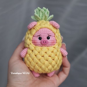 Pode incluir: Um porco de crochê vestindo uma fantasia de abacaxi. O porco é rosa com olhos pretos e um pequeno focinho rosa. O abacaxi é amarelo com folhas verdes.
