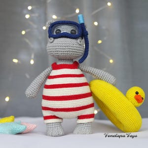 May include: A gray crocheted hippopotamus wearing a red and white striped swimsuit and a blue diving mask. The hippopotamus is holding a yellow crocheted inflatable ring with a yellow duck on top. The background is a blurry image of white lights.