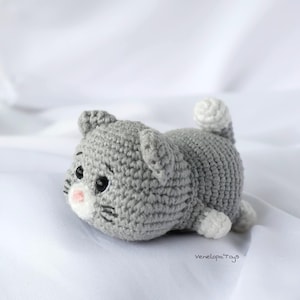 Puede incluir: Un gato de ganchillo hecho a mano en posición tumbada. El gato es principalmente gris con detalles blancos en las patas y la punta de la cola. Tiene ojos negros, bigotes y una nariz rosa. El gato está sobre una superficie blanca.