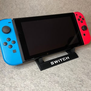 May include: A black Nintendo Switch console with blue and red Joy-Con controllers is on a black stand with the word "SWITCH" printed on it.