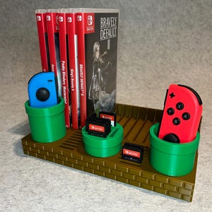 Puede incluir: Un organizador de videojuegos impreso en 3D con temática de Super Mario Bros. Cuenta con soportes verdes con forma de tubería para mandos y cartuchos de juego, y una base marrón con diseño de ladrillos. Se ven varias carcasas de juegos de Nintendo Switch.