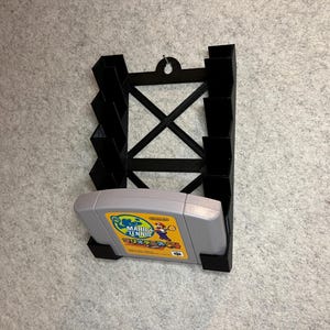 Può includere: Supporto da parete in plastica nera per cartucce di gioco Nintendo 64. Il supporto ha slot per più cartucce e una singola cartuccia è esposta nel supporto. La cartuccia è per il gioco "Mario Tennis".