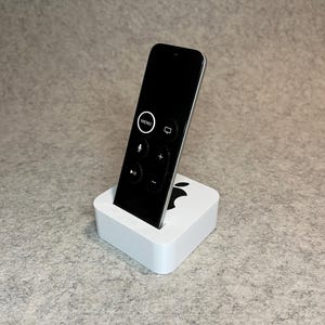 May include: A white charging stand with an Apple logo on the front, holding a black Apple TV remote with a microphone, play, pause, and volume buttons.