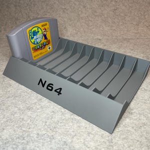 Op de afbeelding: Een grijze Nintendo 64-spelcartridgehouder met een sleuf voor een spelcartridge en acht sleuven voor extra cartridges. De houder heeft de tekst "N64" op de voorkant. Een Nintendo 64-spelcartridge zit in de sleuf, met het spel "Mario Tennis" met een geel en blauw ontwerp.