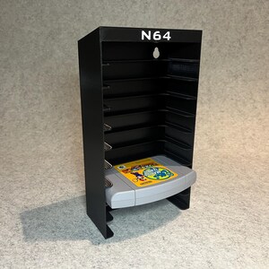 Op de afbeelding: Zwarte N64-videospelcartridgehouder met een grijze cartridge erin. De houder heeft 10 slots voor cartridges en de tekst "N64" aan de voorkant.
