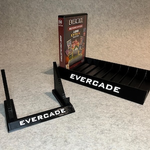Puede incluir: Soporte de plástico negro con la palabra "EVERCADE" impresa en blanco. El soporte está diseñado para sostener una caja de cartucho de videojuego. Una caja de cartucho de plástico negro con un cartucho de videojuego en su interior está sentada en el soporte. La caja de cartucho tiene las palabras "EVERCADE" y "ATARI LYNX COLLECTION 2" impresas en ella.